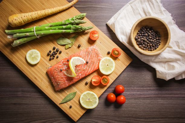 Raw salmon on cutting board with lemon slices asparagus, grape tomatoes, and peppercorns