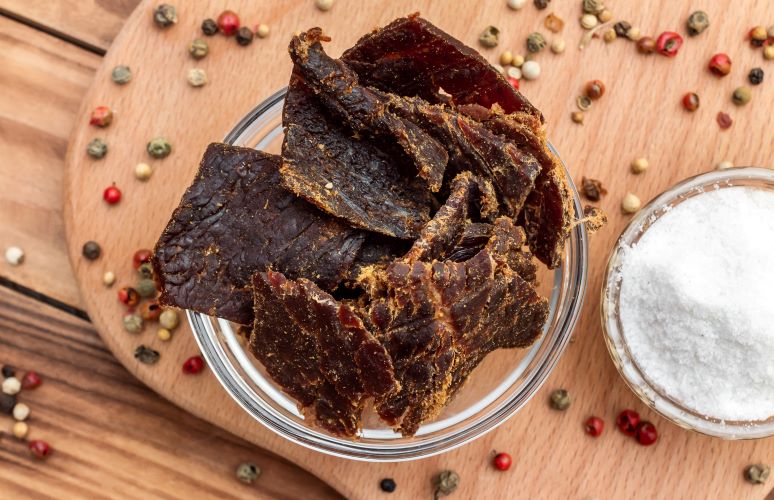 Jerky in a bowl on wood and surrounded by peppercorns