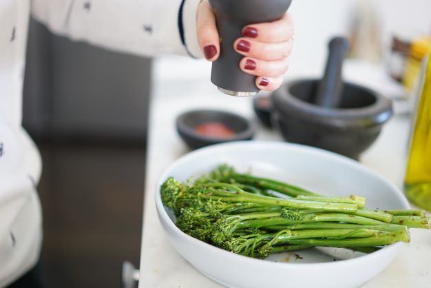 Broccolini in white bowl with a hand crushing black pepper over top