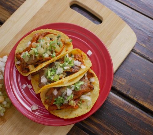 Three corn tacos sitting on a red plate, on top of a cutting board filled with meat and veggies