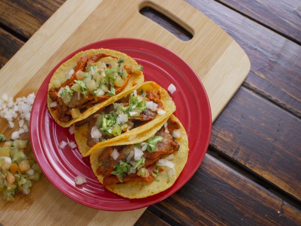 Three corn tacos sitting on a red plate, on top of a cutting board filled with meat and veggies