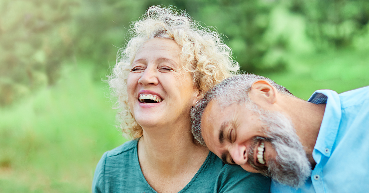 Photograph of a joyful older couple outdoors, with the man leaning his head on the woman's shoulder as they both laugh. This image conveys connection, happiness, and emotional well-being in later life.