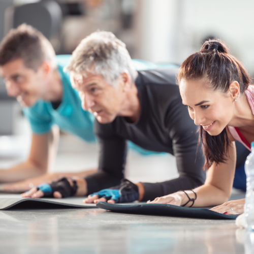 Three people of different ages performing a plank exercise on yoga mats in a gym, smiling and focused during a fitness class