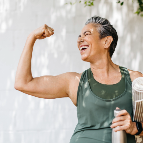 Smiling older adult in workout clothes flexing her arm while holding a yoga mat and water bottle outdoors.