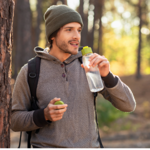 A young man wearing a gray hoodie and green beanie drinks from a reusable water bottle while standing next to a tree in a sunlit forest.