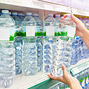 Hands selecting a large plastic water bottle from a supermarket shelf filled with bottled water.