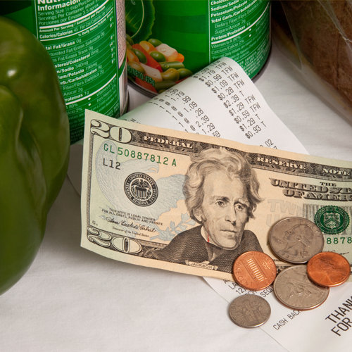 Close-up of grocery receipt with dollar bills and coins placed on top next to fresh vegetables.