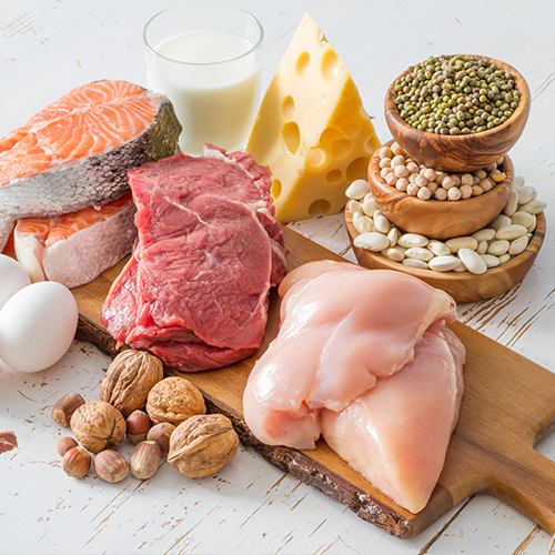 Assortment of protein-rich foods including chicken, beef, salmon, eggs, milk, cheese, beans, nuts, and seeds on a table.