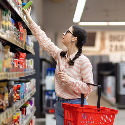 College student with a red basket reaching for an item on the top shelf in a grocery store aisle.